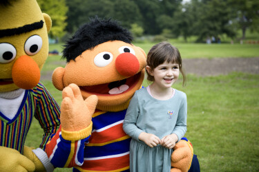 Ernie und Bert winken hinter einem Mädchen in die Kamera.