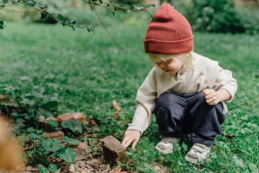 Kind mit roter Mütze hockt auf einer Wiese und erforscht einen Stein.